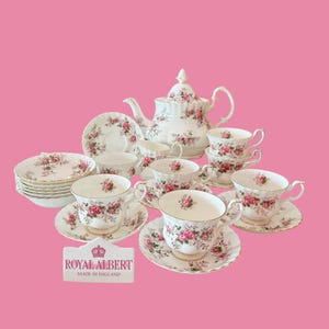Könnte beinhalten: Ein Royal Albert Teeservice bestehend aus einer weißen Porzellanteekanne, einem Milchkännchen und mehreren Teetassen mit passenden Untertassen, verziert mit rosa Rosenmustern und Goldrand. Ein Schild zeigt den Text ROYAL ALBERT MADE IN ENGLAND.