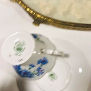 May include: A white teacup and saucer with a floral pattern featuring blue flowers. The teacup has a green mark on the bottom.