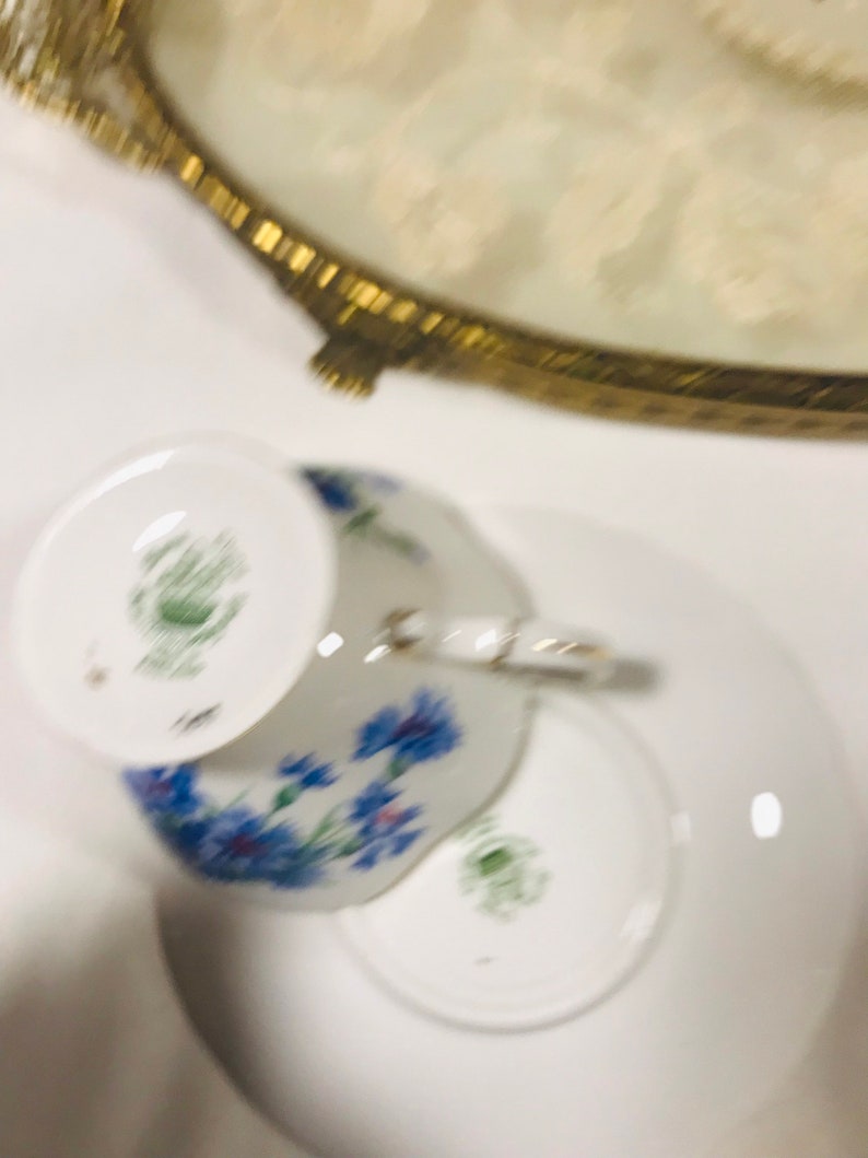 May include: A white teacup and saucer with a floral pattern featuring blue flowers. The teacup has a green mark on the bottom.