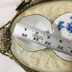 May include: Two white teacups with gold trim and blue floral designs on a gold and white lace tray. A ruler is used to show the size of the teacups, which are approximately 3.5 inches in diameter.