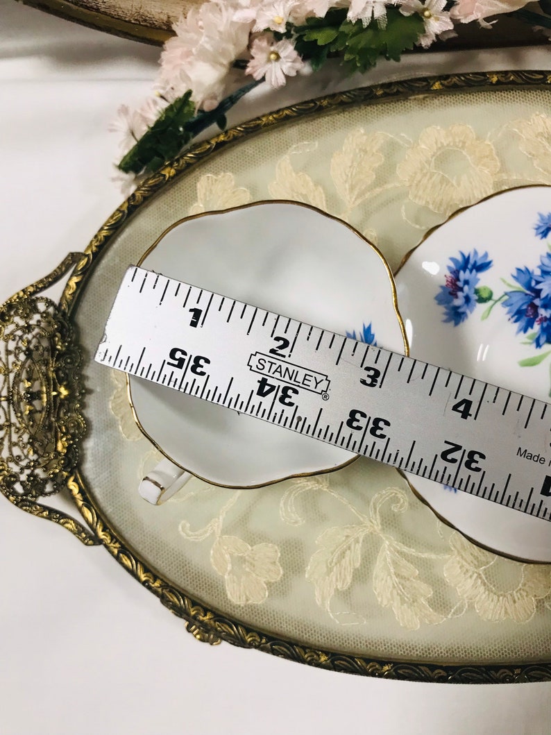 May include: Two white teacups with gold trim and blue floral designs on a gold and white lace tray. A ruler is used to show the size of the teacups, which are approximately 3.5 inches in diameter.