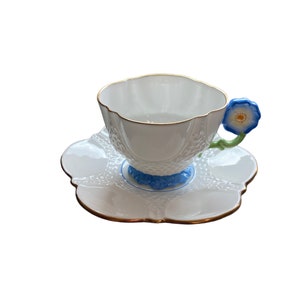 May include: A white teacup and saucer set with a blue flower handle. The cup and saucer have a delicate, embossed design and a gold trim.