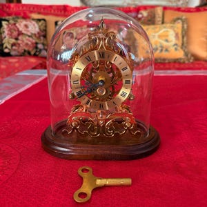 Puede incluir: Un reloj antiguo dorado con números romanos, encerrado en una cúpula de cristal. El reloj se asienta sobre una base de madera, con un diseño ornamentado. Una llave dorada descansa sobre la superficie roja. El reloj mide unos 20 cm de alto.