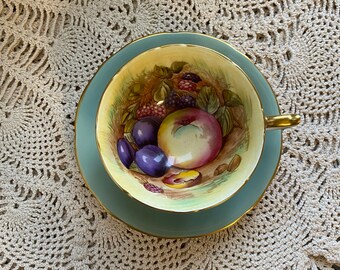 May include: A vintage teacup and saucer with a delicate floral design featuring a peach, plums, and blackberries. The cup and saucer are a pale blue color with a gold trim.
