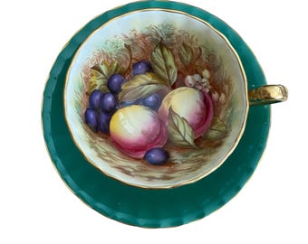 May include: A green teacup and saucer set with a floral design featuring peaches and grapes. The cup and saucer have a gold rim.