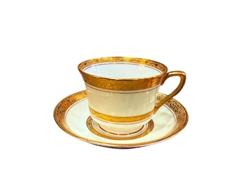 Puede incluir: Una taza de té y un platillo blancos con ribetes dorados. La taza tiene un delicado patrón floral y el platillo tiene una simple banda dorada.