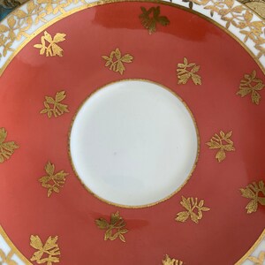 May include: A coral-colored saucer with a white center and a gold rim. The saucer is decorated with a repeating pattern of gold floral motifs.