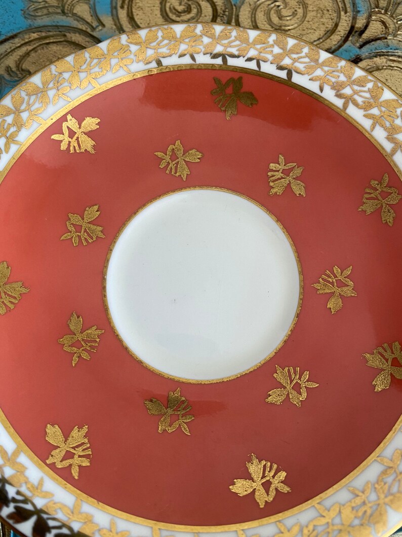 May include: A coral-colored saucer with a white center and a gold rim. The saucer is decorated with a repeating pattern of gold floral motifs.