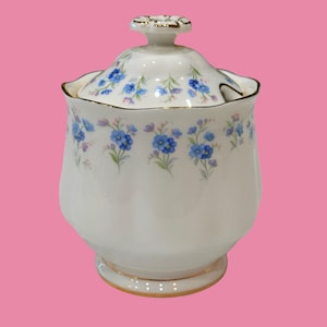 Puede incluir: Un azucarero de porcelana blanca con borde festoneado y tapa a juego con un pomo dorado. El cuerpo y la tapa están decorados con un patrón repetitivo de pequeñas flores nomeolvides azules con tallos y hojas de color verde.