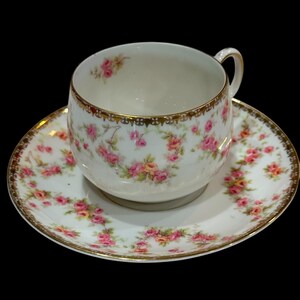 Puede incluir: Un juego de taza y platillo blancos con un estampado floral de rosas rosas y amarillas. Los bordes están adornados con dorado. La taza tiene un asa curvada. El platillo es redondo y tiene un diseño floral a juego. Sobre fondo negro.