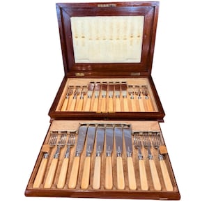 Puede incluir: Una caja de madera con tapa con bisagras, que contiene un juego de cubiertos de plata con mangos de marfil. Los cubiertos están dispuestos en filas, con los cuchillos y los tenedores alternando.
