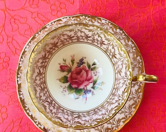 May include: A vintage teacup and saucer set with a delicate floral design. The cup and saucer are white with a gold rim and a pink rose with green leaves in the center. The saucer has a gold floral pattern around the edge.