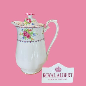 Peut inclure: Pichet en porcelaine blanche avec un motif floral et des bordures dorées. Le pichet a une anse incurvée et un couvercle avec un bouton floral rose. La marque Royal Albert est affichée sur une étiquette blanche.