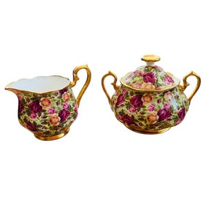 Puede incluir: Un juego de crema y azúcar con un estampado floral. El juego incluye una jarra de crema y un azucarero con tapa, ambos con ribetes y asas doradas. El estampado floral presenta rosas rosas y moradas sobre un fondo crema.