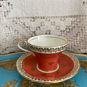 May include: A vintage teacup and saucer set with a coral red base and gold trim. The cup and saucer are decorated with a floral pattern in gold.
