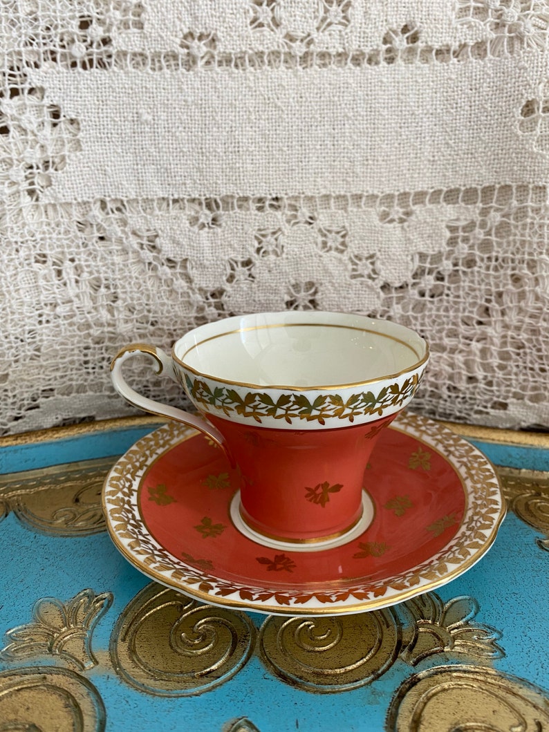 May include: A vintage teacup and saucer set with a coral red base and gold trim. The cup and saucer are decorated with a floral pattern in gold.