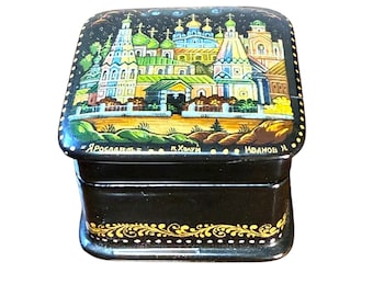 May include: A black lacquered box with a detailed, colorful painting of a cityscape on the lid. The painting features buildings with green, blue, and yellow accents. The box has gold filigree trim and text in Russian.