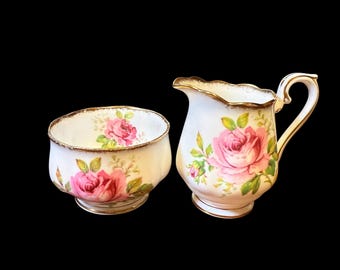 May include: A vintage cream and sugar set. The white porcelain pieces feature pink rose floral designs with green leaves and gold trim. The sugar bowl is round, and the creamer has a handle and spout.