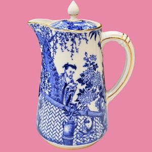 Peut inclure: Une cruche en porcelaine blanche avec un motif bleu de type transfert représentant des personnages, des arbres et des fleurs. La cruche comprend un couvercle assorti avec un bouton, une anse courbée et un liseré doré sur le bord.