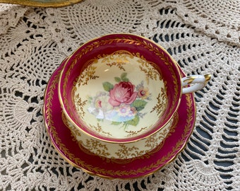 May include: A vintage teacup and saucer set with a floral design. The cup and saucer are both decorated with a gold trim and a burgundy background. The floral design features a pink rose and other flowers in shades of pink, yellow, and white.