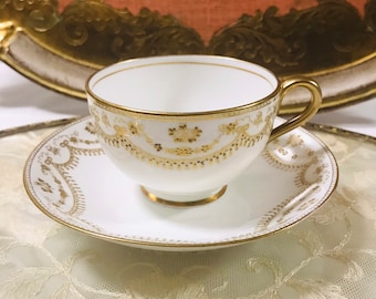 Puede incluir: Una taza de té y un platillo blancos con ribete dorado y un delicado patrón floral. La taza y el platillo están sobre un encaje blanco.