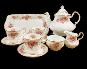 Könnte beinhalten: Ein weißes Teeservice mit Blumenmuster. Das Set beinhaltet eine Teekanne, eine Milchkännchen, eine Zuckerdose und vier Teetassen mit Untertassen. Das Blumenmuster zeigt rosa Rosen auf weißem Hintergrund.