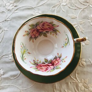 May include: A white teacup and saucer with a green rim and gold trim. The teacup features a floral design with pink roses and green leaves.
