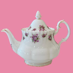 May include: A white porcelain teapot featuring a scalloped body and a matching lid with a pointed finial. The teapot is decorated with small clusters of purple and white flowers and green leaves, accented with gold trim on the edges and handle.