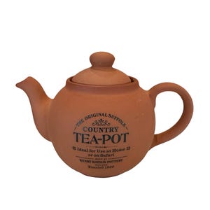 May include: A terracotta-colored teapot with a rounded body, spout, and handle. The lid has a small knob. The text "COUNTRY TEA-POT" is printed on the side, along with additional text. The teapot is designed for home or safari use.