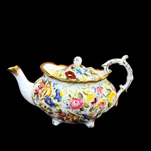 May include: A white teapot with a floral pattern and gold accents. The teapot has a rounded body and a curved spout. The floral pattern features a variety of flowers in shades of pink, yellow, blue, and red.