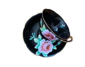 Puede incluir: Juego de taza y platillo de té negro con borde dorado y diseño floral de rosas rosas y verdes. La taza está inclinada, revelando su interior. El platillo tiene un borde festoneado.