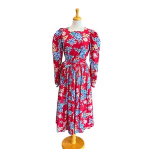 May include: A red floral print dress with long sleeves and a square neckline. The dress features a blue and white floral pattern and is displayed on a wooden dress form. The dress has a fitted waist and a flowing skirt.