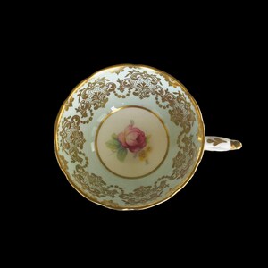 May include: A vintage teacup with a delicate floral design and gold trim. The cup is light blue with a white centre featuring a pink rose and green leaves. The gold trim is intricate and detailed.