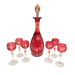 Könnte beinhalten: Ein rubinrotes Kristall-Dekanter-Set mit passendem Stopfen und sechs Weingläsern. Die Karaffe und die Gläser weisen ein geschliffenes Design auf. Das Set wird auf weißem Hintergrund präsentiert.