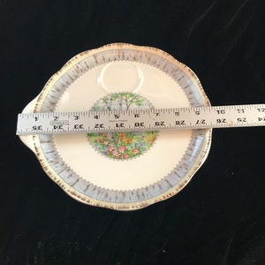 Pu&ograve; includere: Un piatto da portata rotondo, bianco sporco, con bordo smerlato e manico. Il piatto presenta un bordo blu e oro e un'immagine centrale di una scena di giardino. Il piatto misura circa 30 cm di diametro.