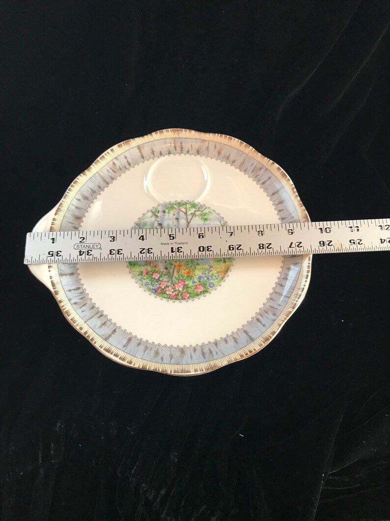 Pu&ograve; includere: Un piatto da portata rotondo, bianco sporco, con bordo smerlato e manico. Il piatto presenta un bordo blu e oro e un'immagine centrale di una scena di giardino. Il piatto misura circa 30 cm di diametro.