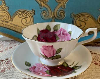 May include: A white teacup and saucer with a pink and red rose pattern. The cup and saucer have a gold trim.