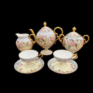 May include: A vintage tea set comprising a teapot, sugar bowl, creamer, and two teacups with saucers. The set has a white and light blue base, pink rose floral patterns, and gold detailing.