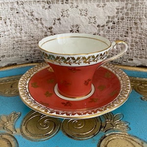 May include: A vintage teacup and saucer set with a coral red base and gold trim. The cup and saucer are decorated with a delicate floral pattern in gold.