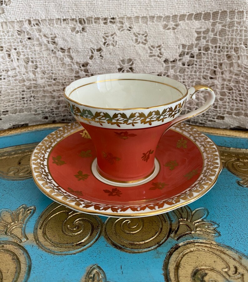 May include: A vintage teacup and saucer set with a coral red base and gold trim. The cup and saucer are decorated with a delicate floral pattern in gold.