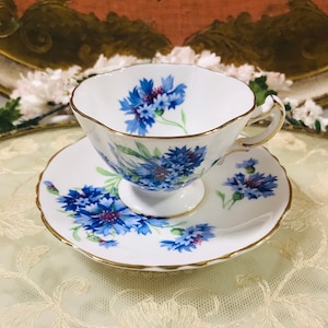 May include: A white teacup and saucer with a blue and gold floral pattern. The flowers are blue cornflowers with green stems and leaves. The cup and saucer have a gold trim.