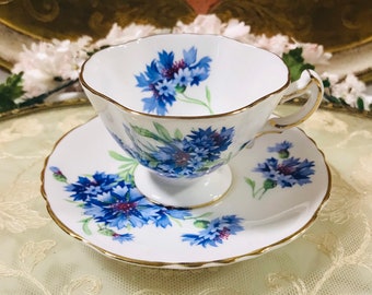 May include: A white teacup and saucer with a blue and gold floral pattern. The flowers are blue cornflowers with green stems and leaves. The cup and saucer have a gold trim.