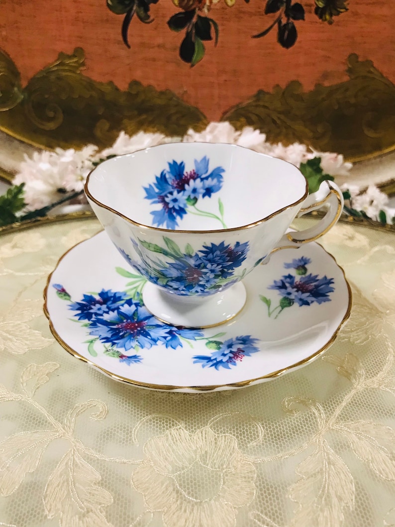 May include: A white teacup and saucer with a blue and gold floral pattern. The flowers are blue cornflowers with green stems and leaves. The cup and saucer have a gold trim.