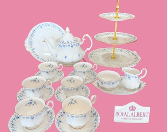 May include: A Royal Albert tea set with a blue floral design. The set includes teacups, saucers, a teapot, a serving plate, a creamer, a sugar bowl, and a three-tiered serving tray. The Royal Albert logo is visible.