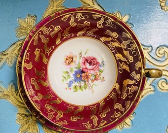 May include: A vintage teacup and saucer set. The cup and saucer are burgundy with gold filigree and a floral center. The cup has a gold handle. The set is on a light blue surface with gold decorative accents.