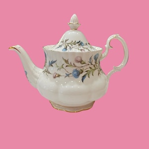 Puede incluir: Una tetera de porcelana blanca con base festoneada y tapa a juego con remate puntiagudo. El cuerpo está decorado con un patrón botánico de flores de cardo azules y rosas, hojas verdes y tallos marrones finos, acentuado con bordes dorados.