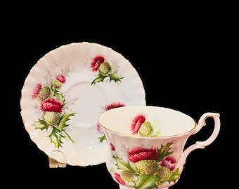 May include: A white teacup and saucer set with a floral design featuring pink and green thistles. The set is made of fine china and has a delicate gold trim.