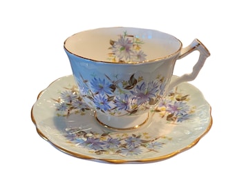 Könnte beinhalten: Ein zartes Tee-Set mit Blumenmuster. Tasse und Untertasse sind weiß mit hellblauem Hintergrund und zeigen ein Blumenmuster mit blauen, violetten und grünen Blüten. Die Ränder von Tasse und Untertasse sind mit Gold verziert.