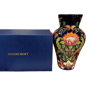 May include: A decorative vase with a dark blue background and a floral design in orange, pink, green, and yellow. The vase is next to a dark blue box with the word "MOORCROFT" in gold lettering.
