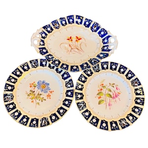 Puede incluir: Juego de tres platos de porcelana vintage con ribete azul y dorado. Los platos presentan diseños florales pintados a mano en tonos rosa, azul y amarillo.